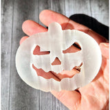 Selenite pumpkin carving ornament held in hand; translucent white disc with jack-o'-lantern face cutouts.