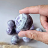 Dream Amethyst sphere, 3.5–4.5 cm, polished with lavender-white banding; held in hand with amethyst spheres visible.