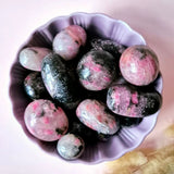 Moonlight Gemstones rhodonite palm stones, tumbled 3–5 cm, pink and black polished stones in a purple bowl