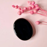 Zwarte obsidiaan duimsteen, Moonlight Gemstones; glanzende ovale steen op roze achtergrond met roze bloemtakjes.