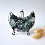 Moss agate moon crescent on black metal display stand; polished druzy textures in green, white and dark tones.